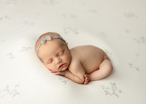 Peaceful newborn baby girl sleeping curled up on white floral blanket wearing a delicate gray bow headband.