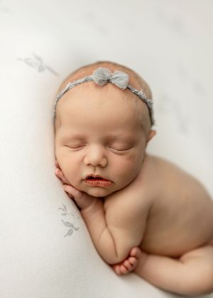 Sleeping newborn baby girl wearing a delicate gray bow headband, peacefully resting on a soft white backdrop.