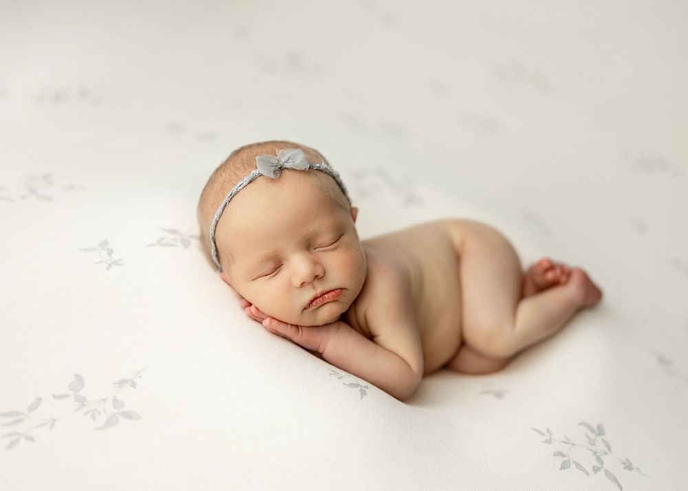 Peaceful newborn baby sleeping on a white floral blanket wearing a delicate gray headband in a professional photo.