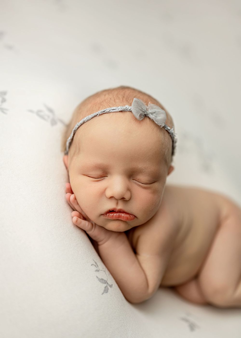 Peaceful sleeping newborn baby girl wearing a delicate gray bow headband, resting on a soft white floral blanket.
