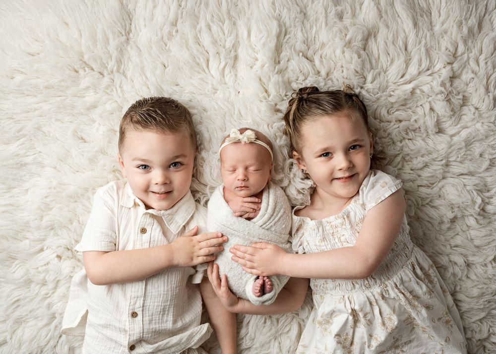 Two children in white outfits lovingly embrace a newborn baby on a soft cream-colored blanket.