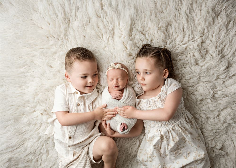 Three children in white outfits lying together on a plush white fur rug during a newborn photo session.
