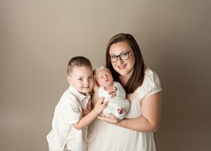 Family studio portrait with young boy and newborn baby against neutral beige background.