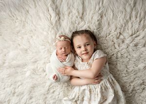 Siblings wearing white dresses pose together on a plush cream-colored fur blanket for a professional portrait.