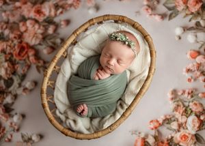 A sleeping newborn swaddled in sage green fabric lies in a rattan basket surrounded by peachy pink flower petals.