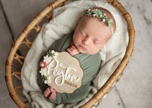 Newborn baby swaddled in sage green wrap sleeping peacefully in round wooden basket with floral headband and wooden sign.
