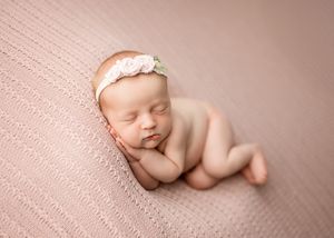 Peacefully sleeping newborn baby curled up on soft pink blanket wearing delicate headband in studio portrait.