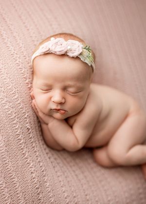 Sleeping newborn on pink knitted blanket wearing delicate floral headband in soft natural light.