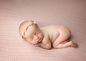 Sleeping newborn wearing delicate floral headband curled up peacefully on soft pink textured blanket.
