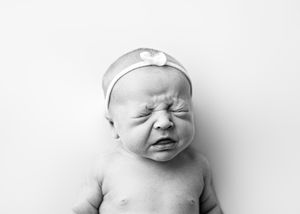A black and white portrait of a crying newborn baby wearing a headband against a plain white background.