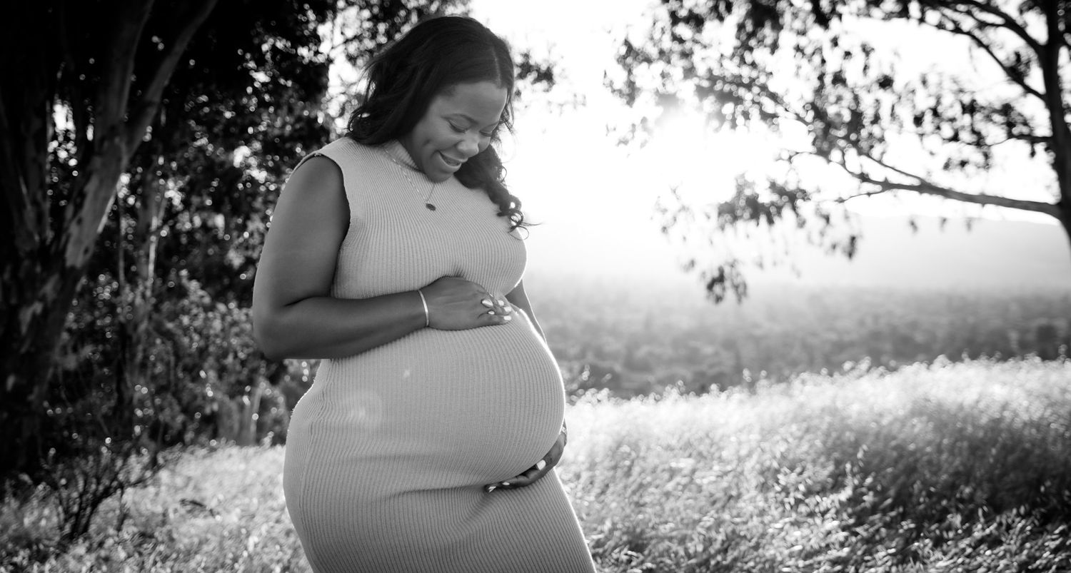 Black and white silhouette of an expectant woman in a sundress touching her belly in a serene outdoor setting at sunset.