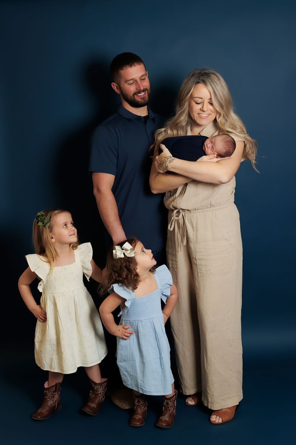A family of five poses for a studio portrait against a dark blue background wearing coordinated neutral colors.