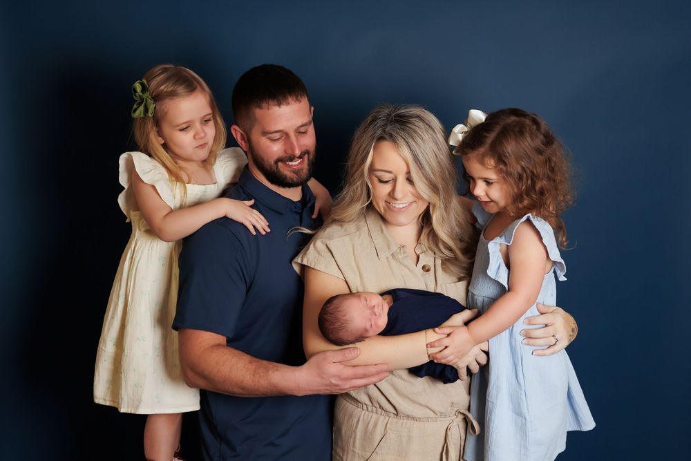 A family with parents wearing navy and beige holds a newborn while two young girls in dresses stand close by.