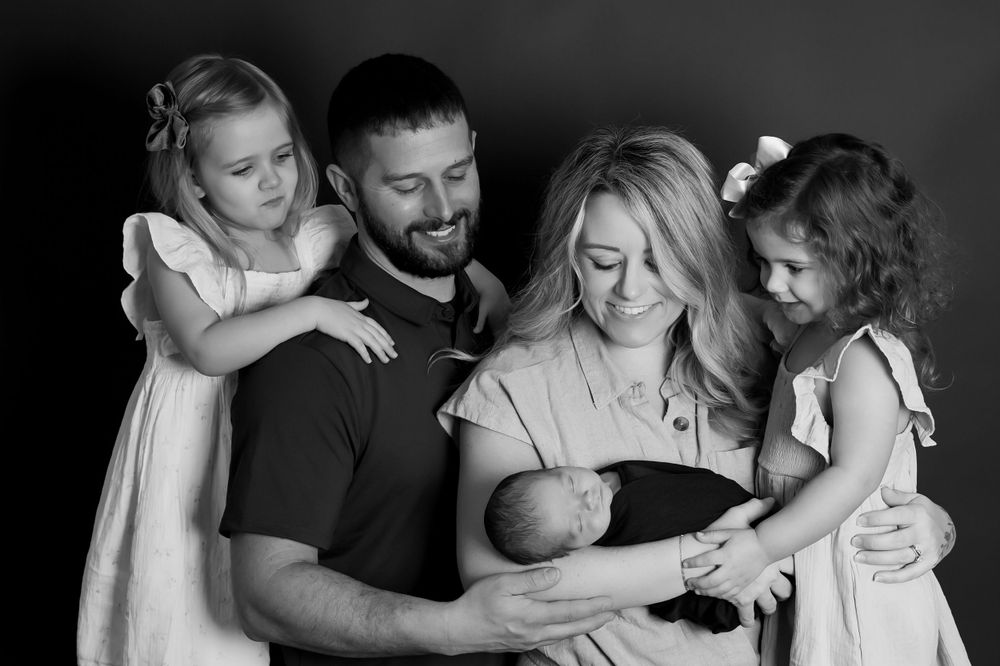 A black and white intimate family portrait showing a group gathered around a newborn baby in a tender moment.