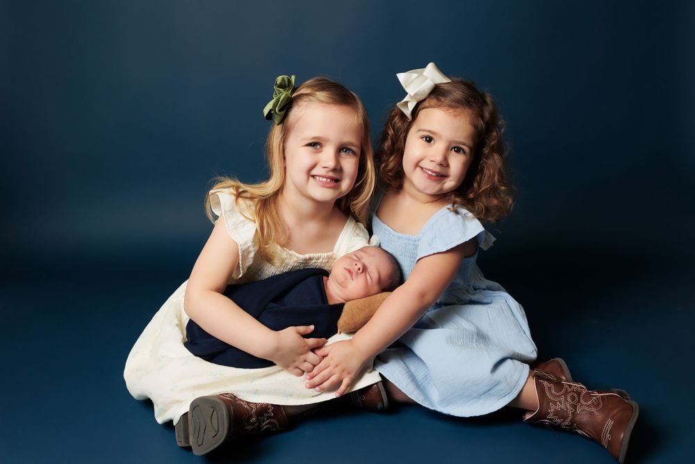 Two young children in dresses sitting together on floor against dark blue backdrop.