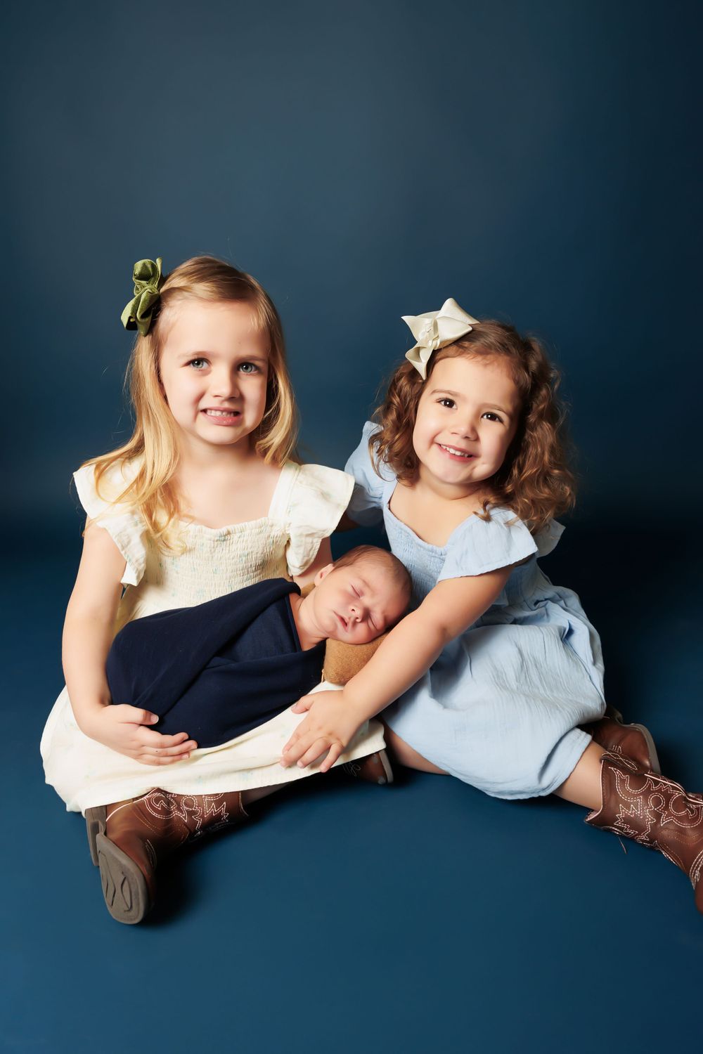 Two young sisters in dresses and cowboy boots sitting together with baby sibling on dark blue background.