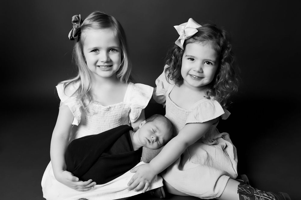 A black and white portrait of two young siblings with a baby sitting together in formal attire.