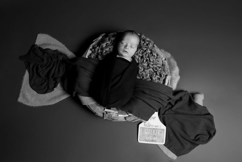 A sleeping newborn lies peacefully wrapped in dark fabric against a textured background in black and white.