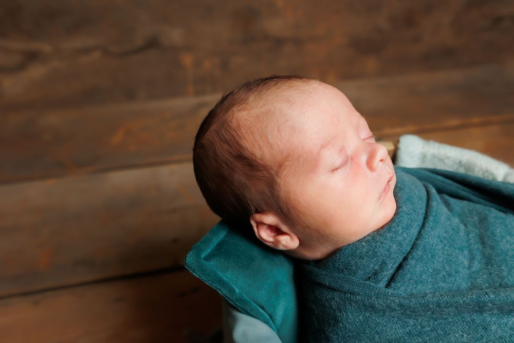Peaceful newborn baby sleeping wrapped in teal swaddle against wooden background.