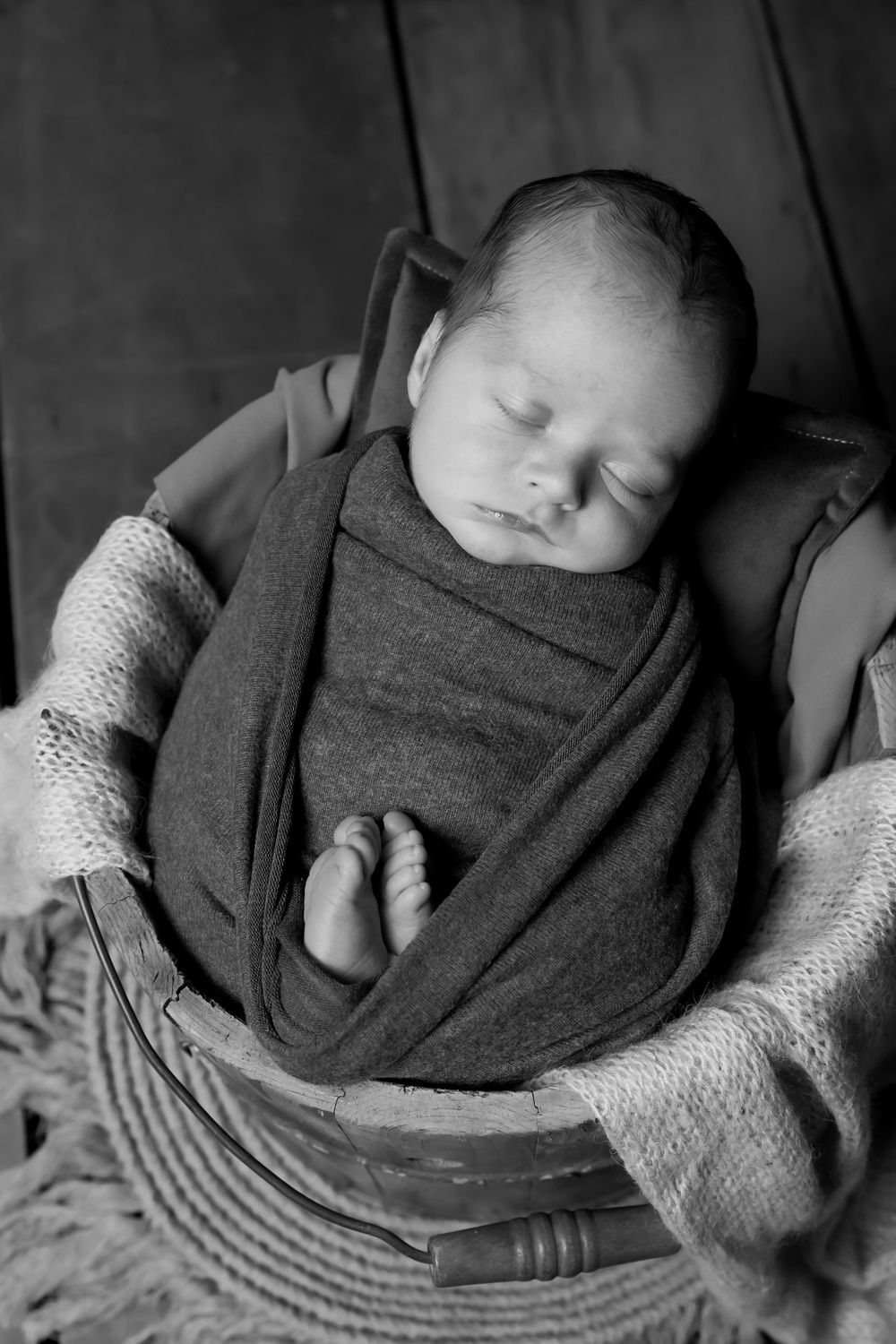 Black and white portrait of sleeping newborn in wooden bucket with textured blankets.