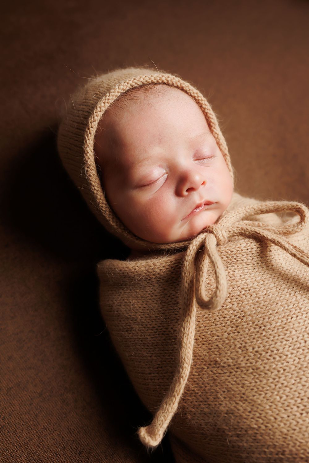 A sleeping newborn baby wrapped in a tan knit bonnet and swaddle on a brown backdrop.