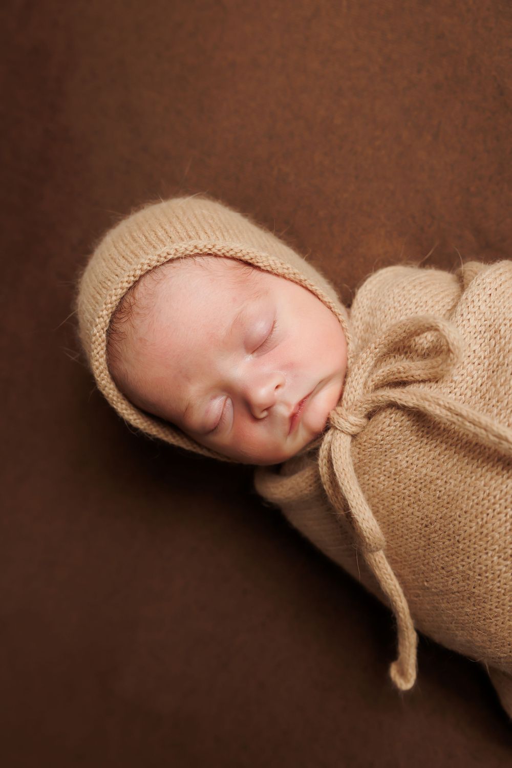Sleeping newborn wrapped in beige knit blanket and bonnet against brown background.