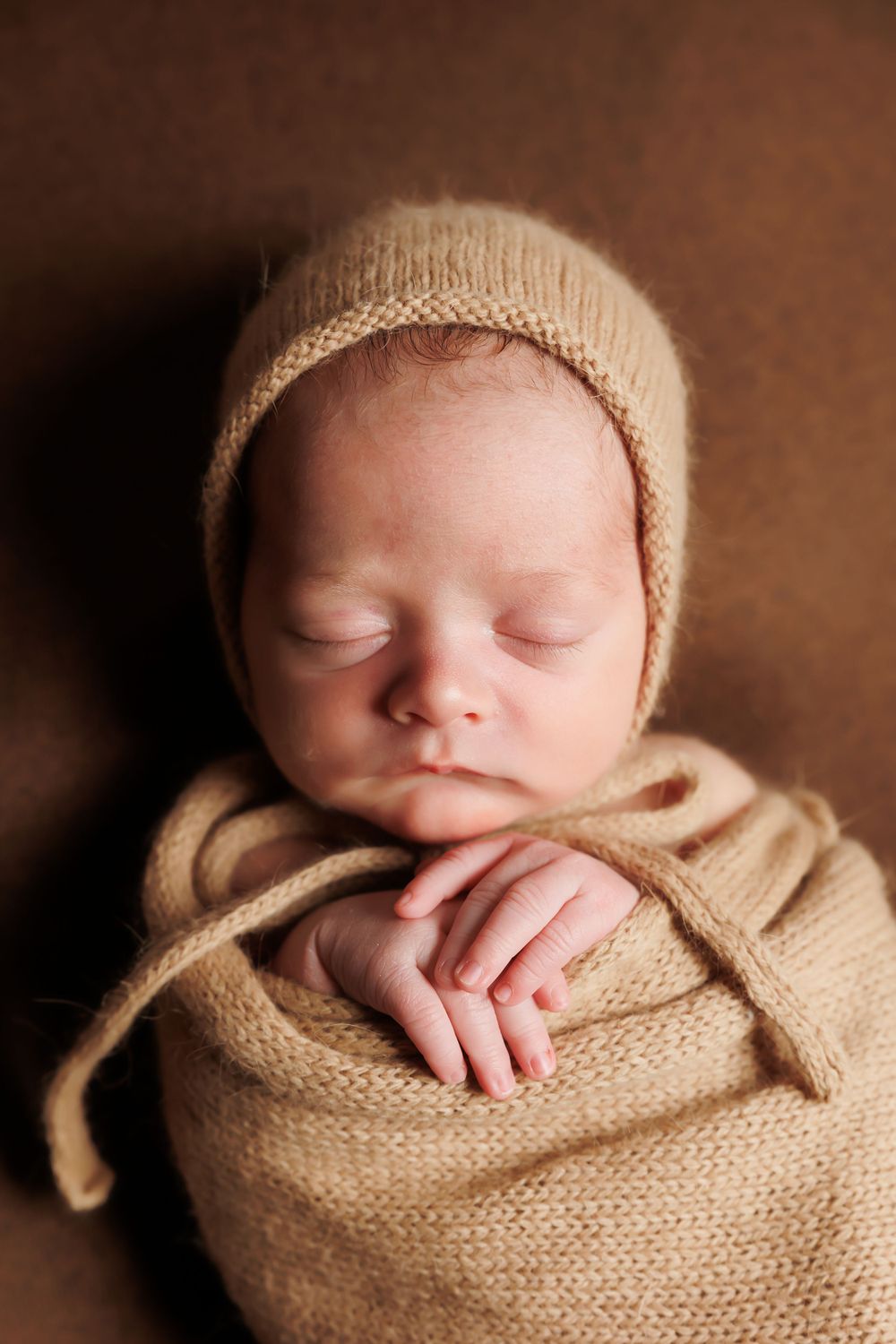 Peaceful newborn baby in beige knit bonnet and wrap against warm brown backdrop.
