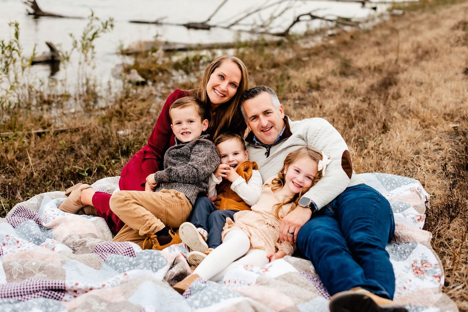 Group relaxes together on cozy blankets during outdoor photo session near water on autumn hillside.