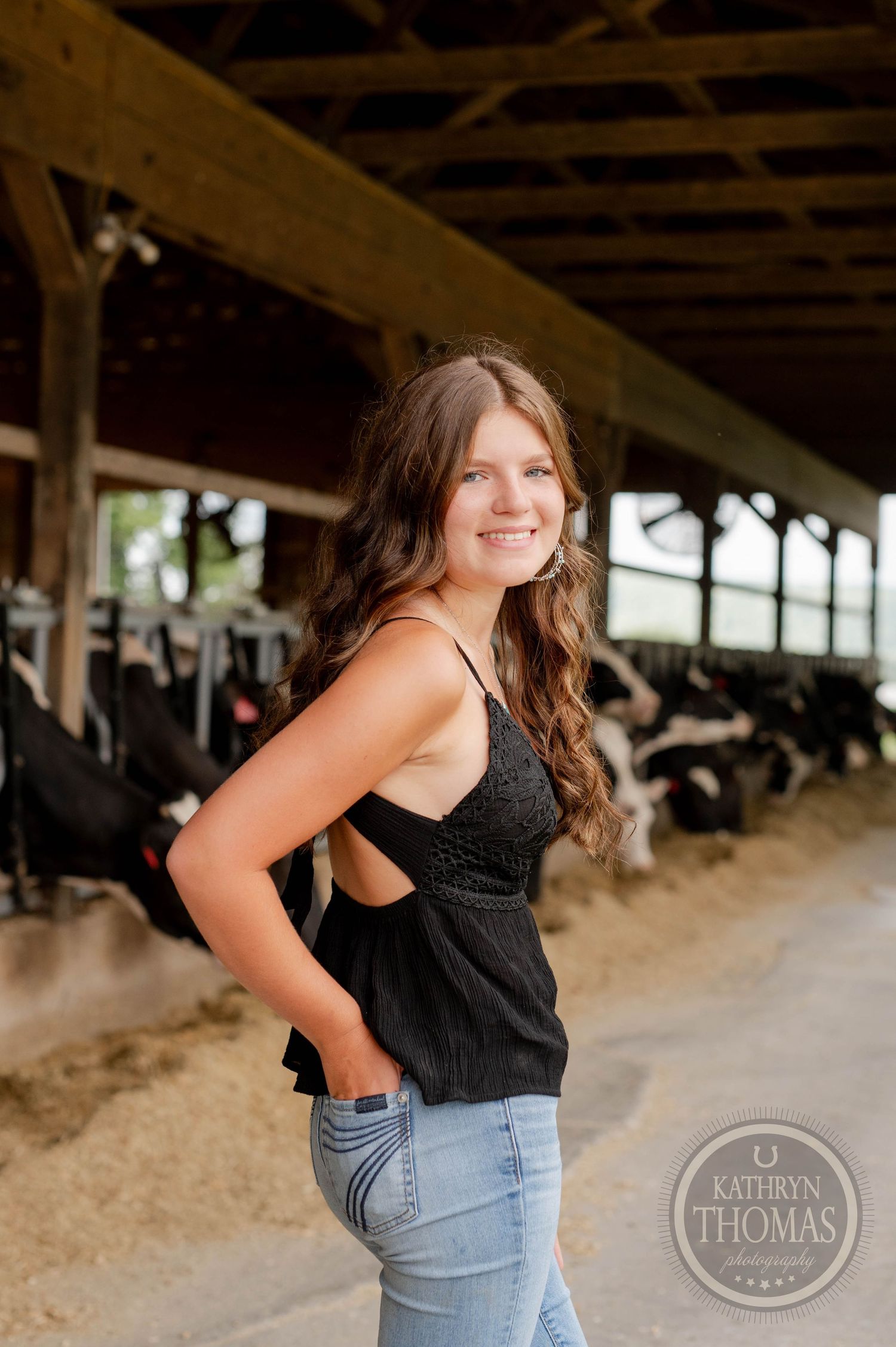 Long-haired person in black tank top poses in dairy barn with Holstein cows visible in background.