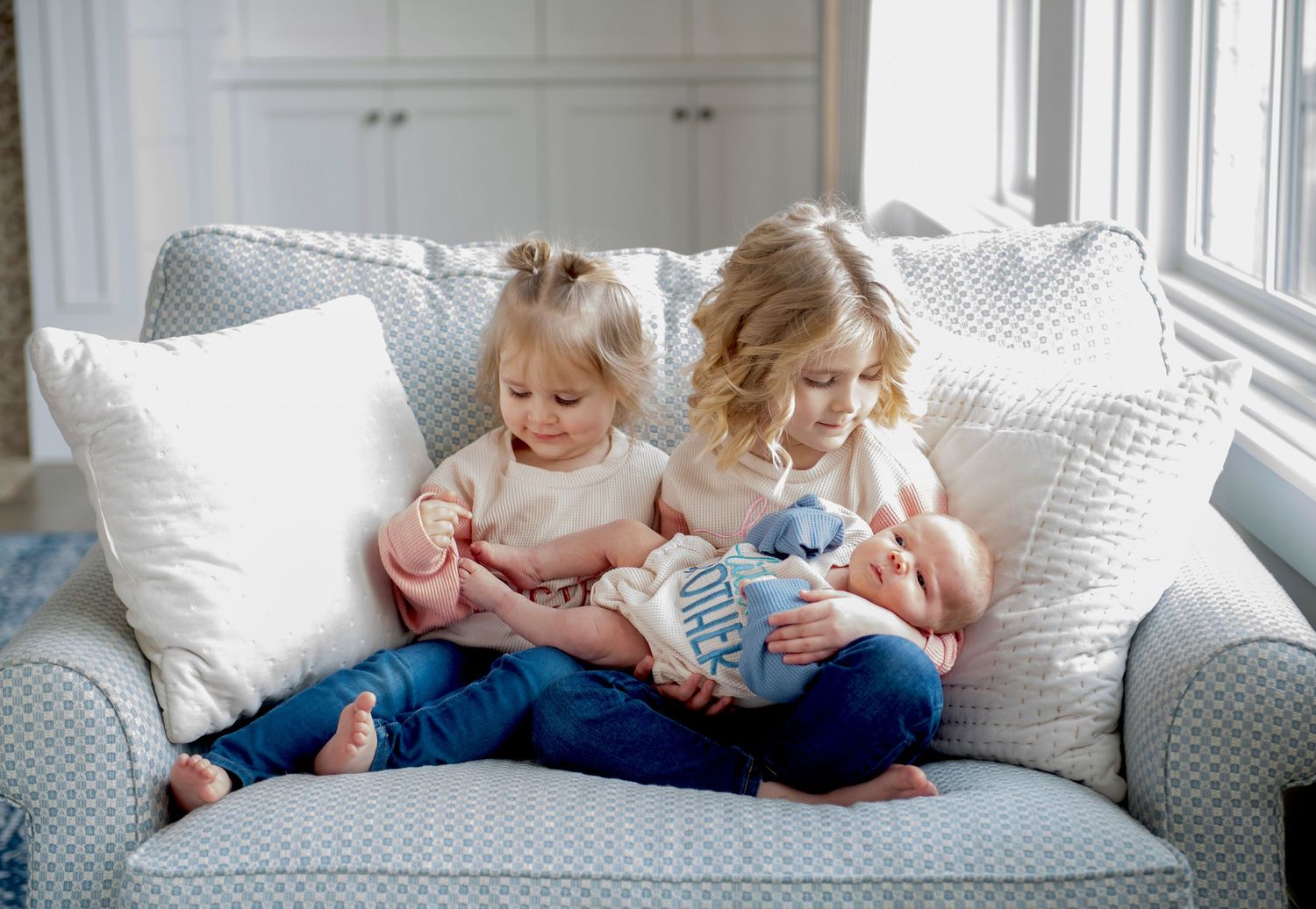 Two small children sitting together on a cozy gray couch with white pillows while looking at electronic devices.