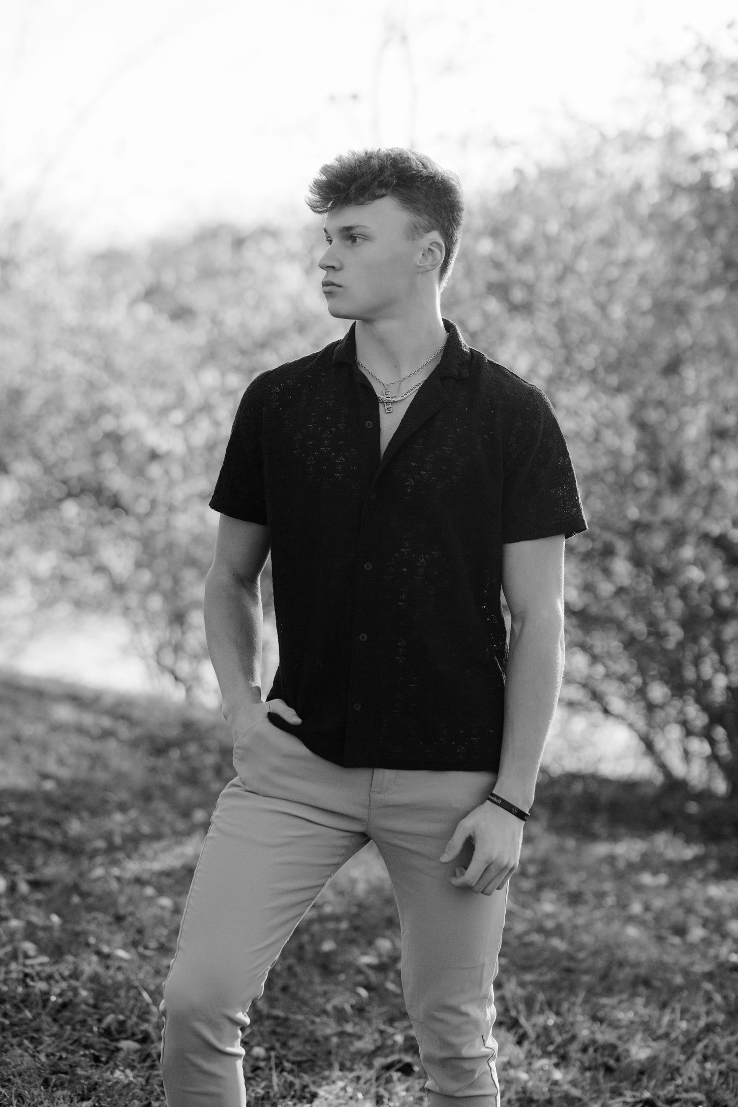 Black and white portrait of person in black polo shirt and light pants standing outdoors against natural background.