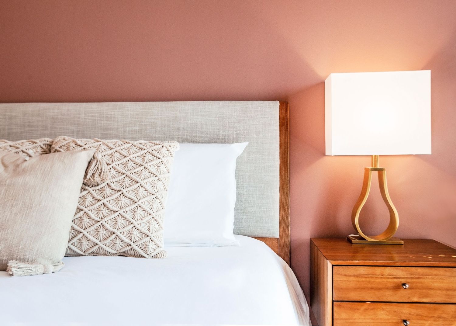 A cozy bedroom interior with pink walls, white bedding, textured throw pillows, and a wooden nightstand with table lamp.