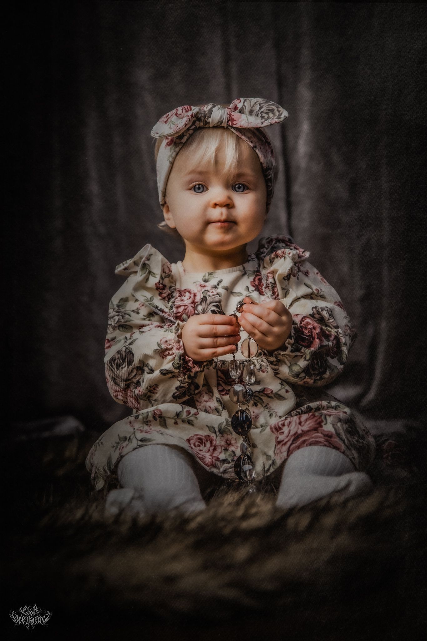 A baby in a floral dress and headband sitting on a dark textured surface.