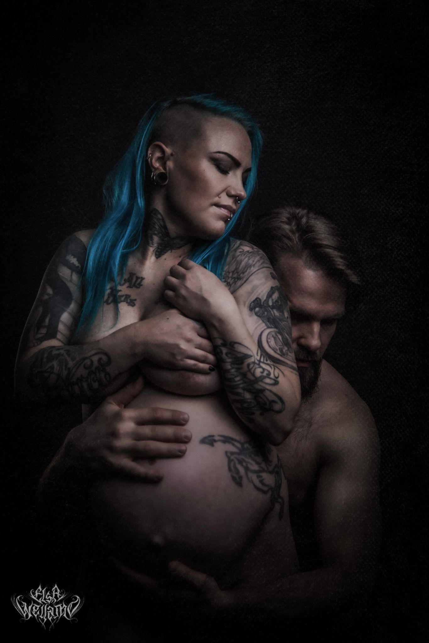 Intimate maternity portrait with blue-haired tattooed woman embraced in dark moody lighting.