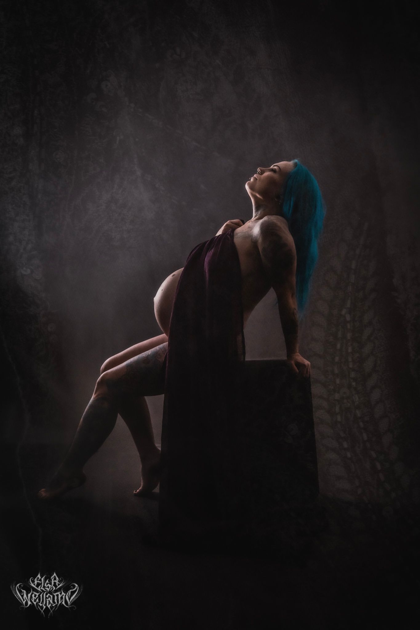 Dramatic low-key portrait of a figure with blue hair posing on dark block against textured wall in moody lighting.