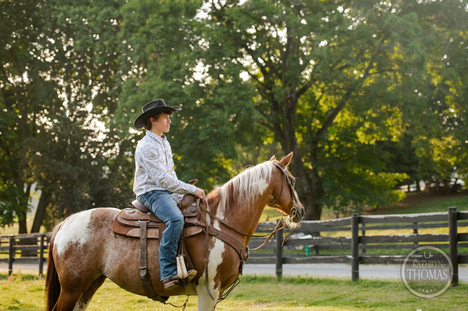 A person rides a paint horse in western tack near a wooden fence at sunset.