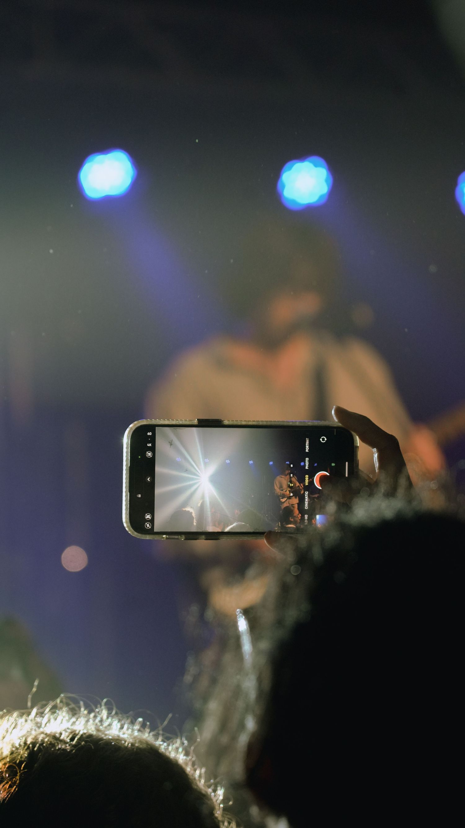 A hand holds an illuminated smartphone displaying video content in a dark setting with blurred lights in the background.