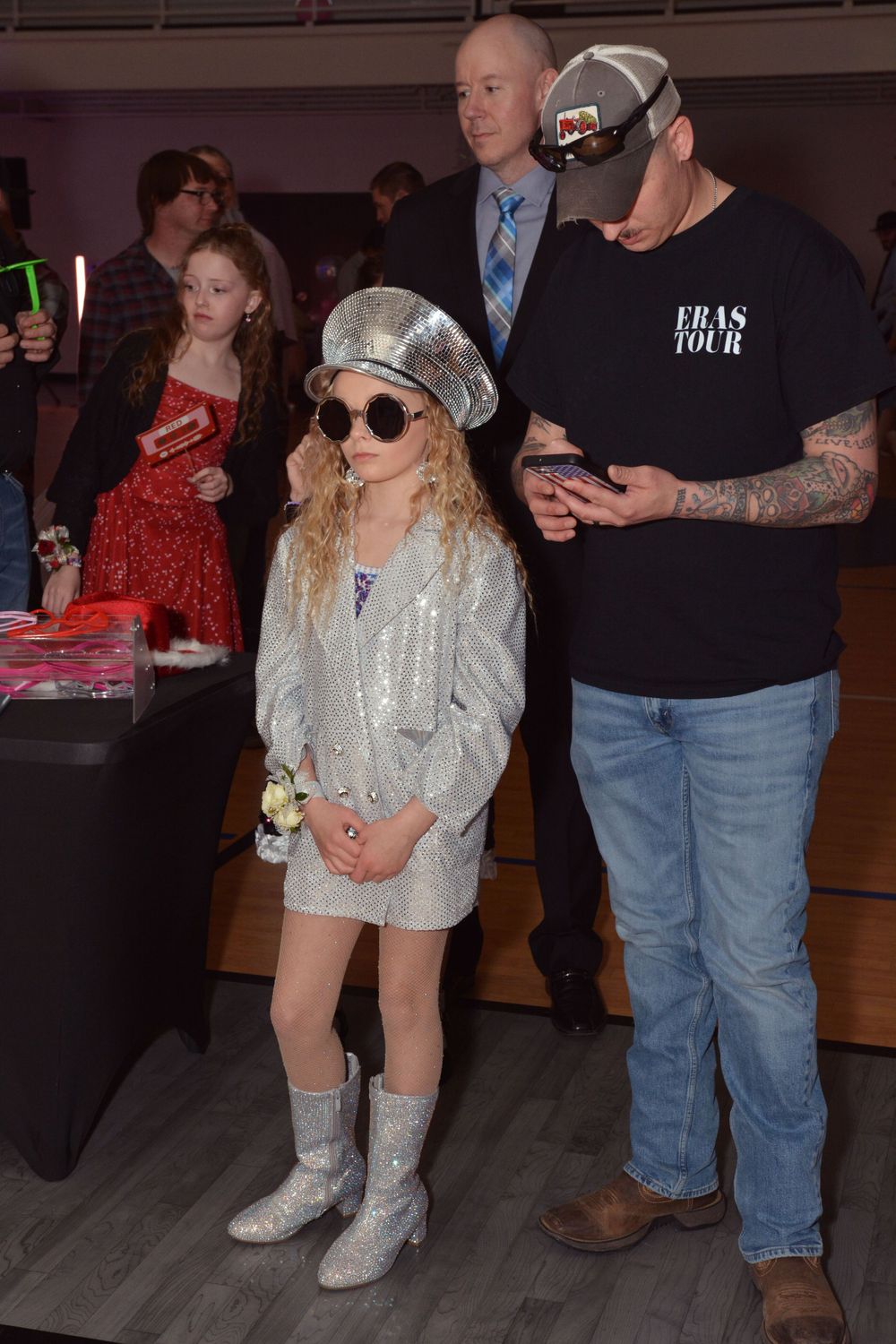 A young girl wearing a sparkly silver dress, sequined cap and boots stands in a casual indoor setting during an event.