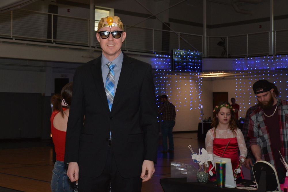 Person in black suit and blue tie wearing sunglasses stands in front of decorative blue lights at indoor event.