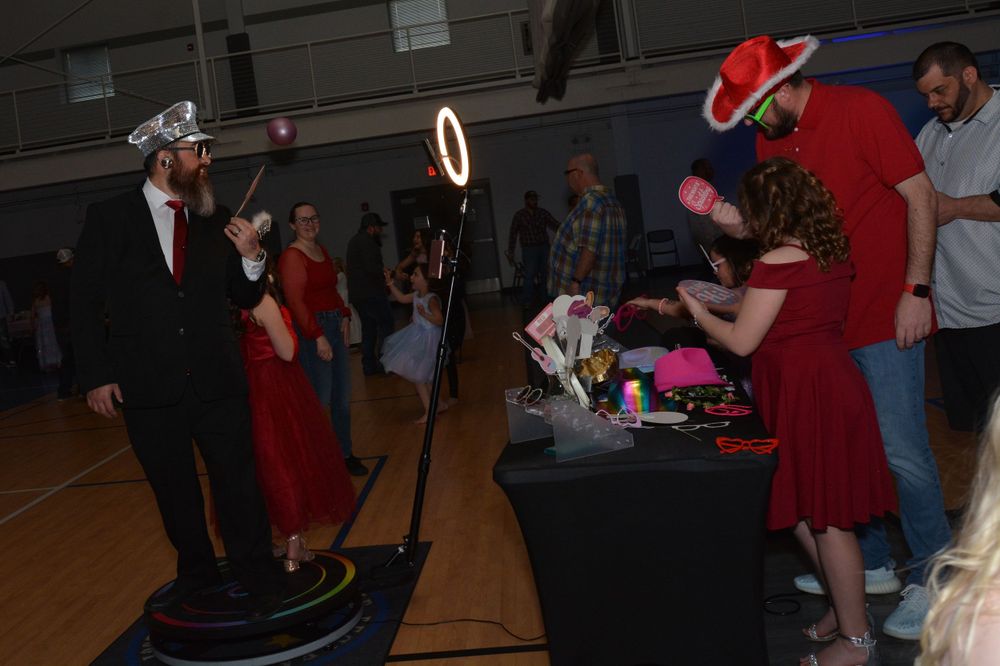People stand around DJ equipment and lighting at an indoor event in a gymnasium space.