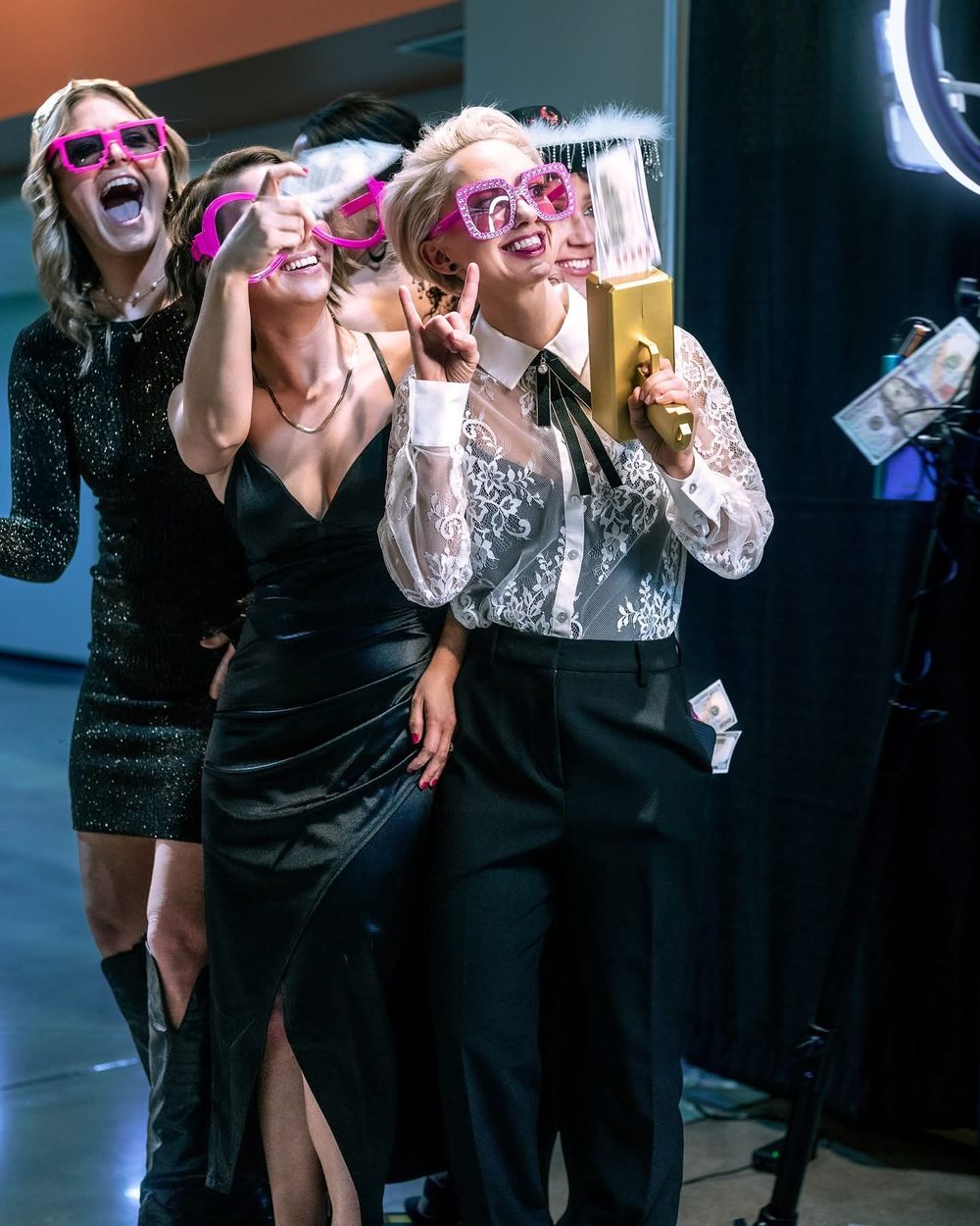 Three friends in formal attire laugh and celebrate with drinks at a party wearing pink novelty glasses.