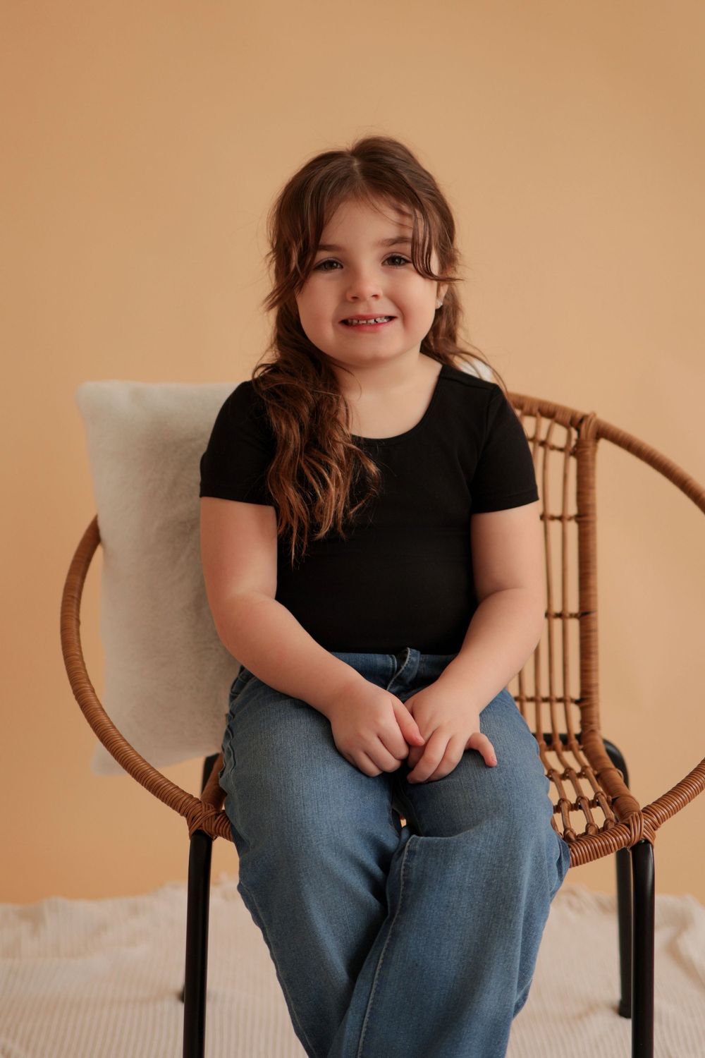 A child sits on a wooden chair against a warm beige background wearing casual clothes.