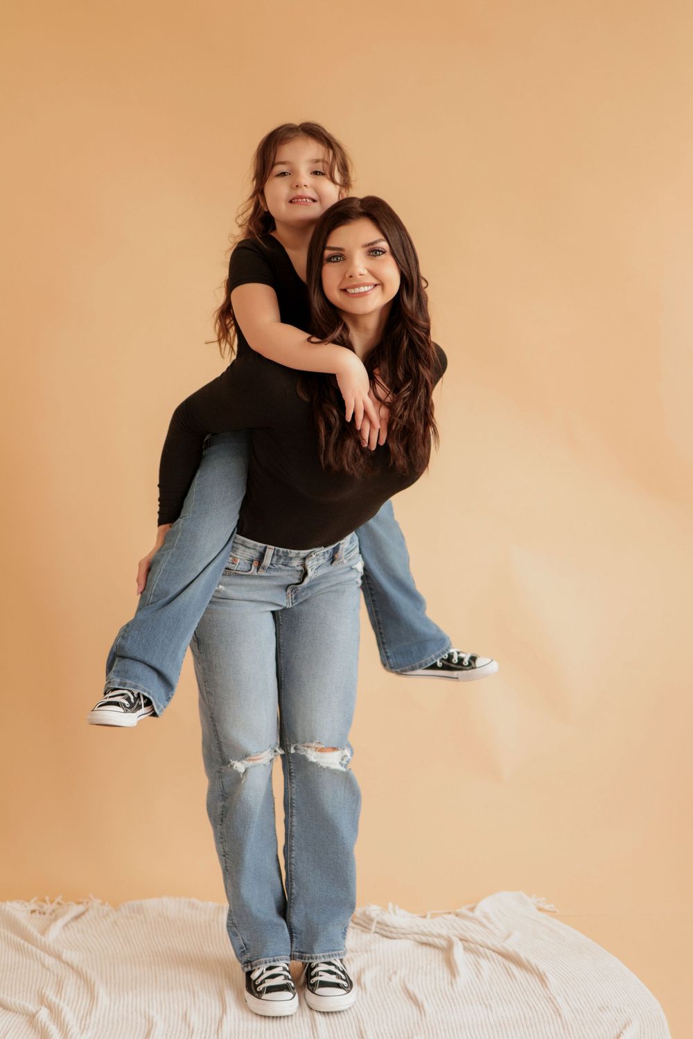Woman in black shirt and ripped jeans poses playfully against beige backdrop.