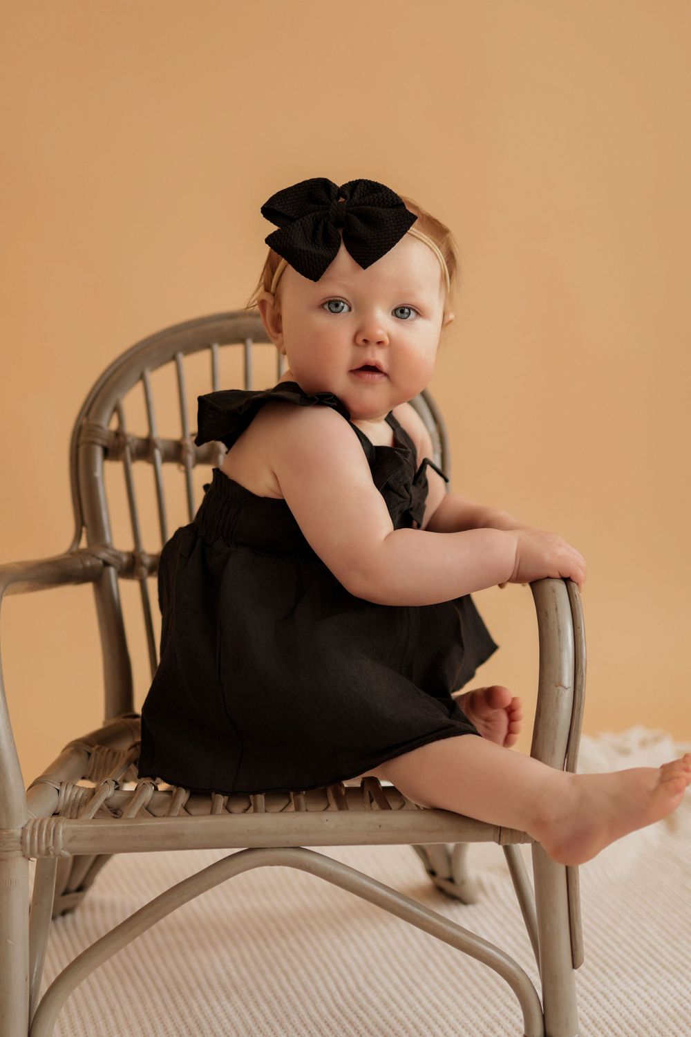 Baby in black dress poses on small gray chair during studio photoshoot.