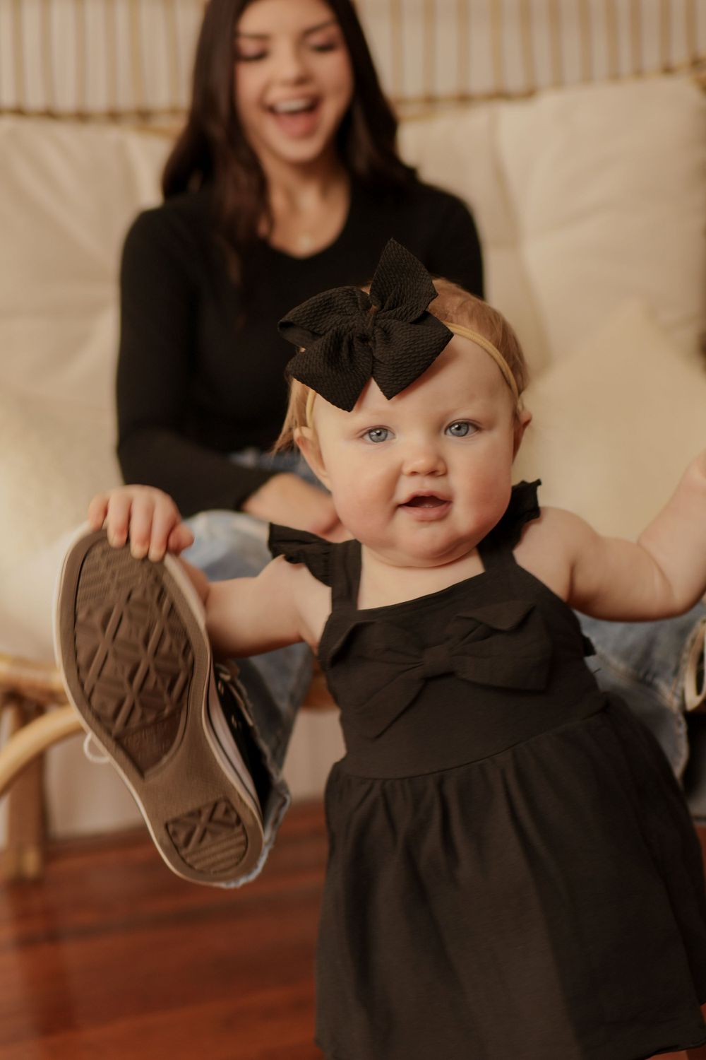 Sweet candid moments captured of small child in black dress showing off shoes on hardwood floor.
