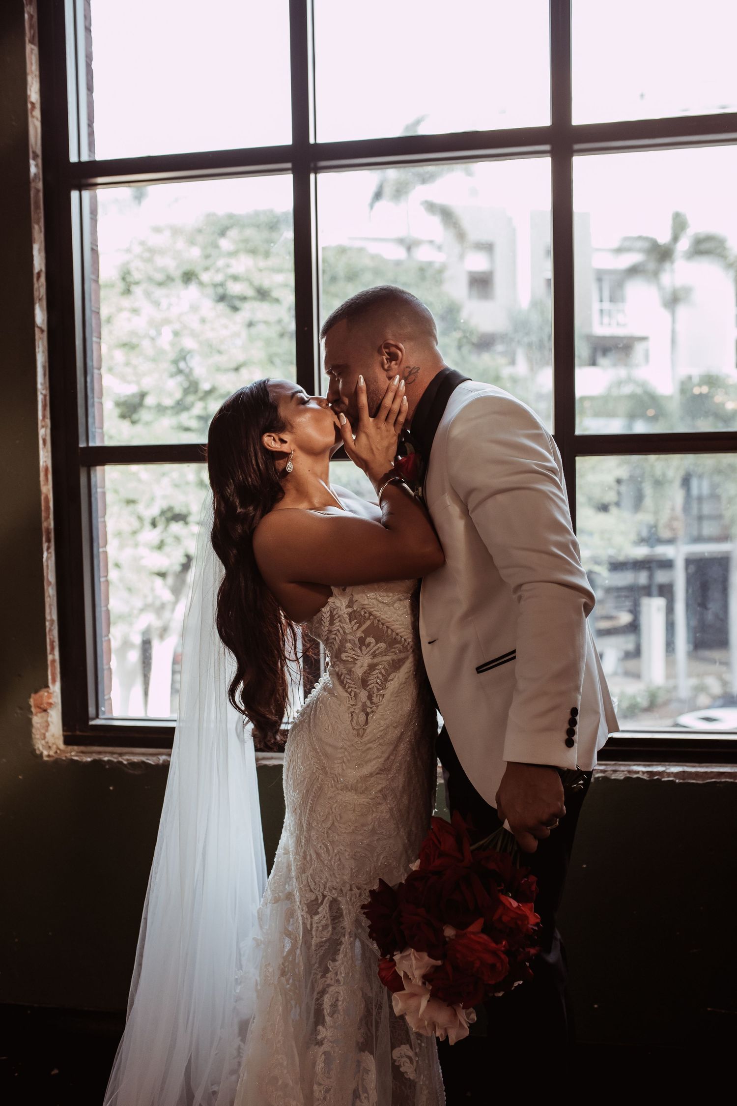 A tender wedding moment between a couple by a window, with the bride in a lace gown and groom in tan suit embracing.