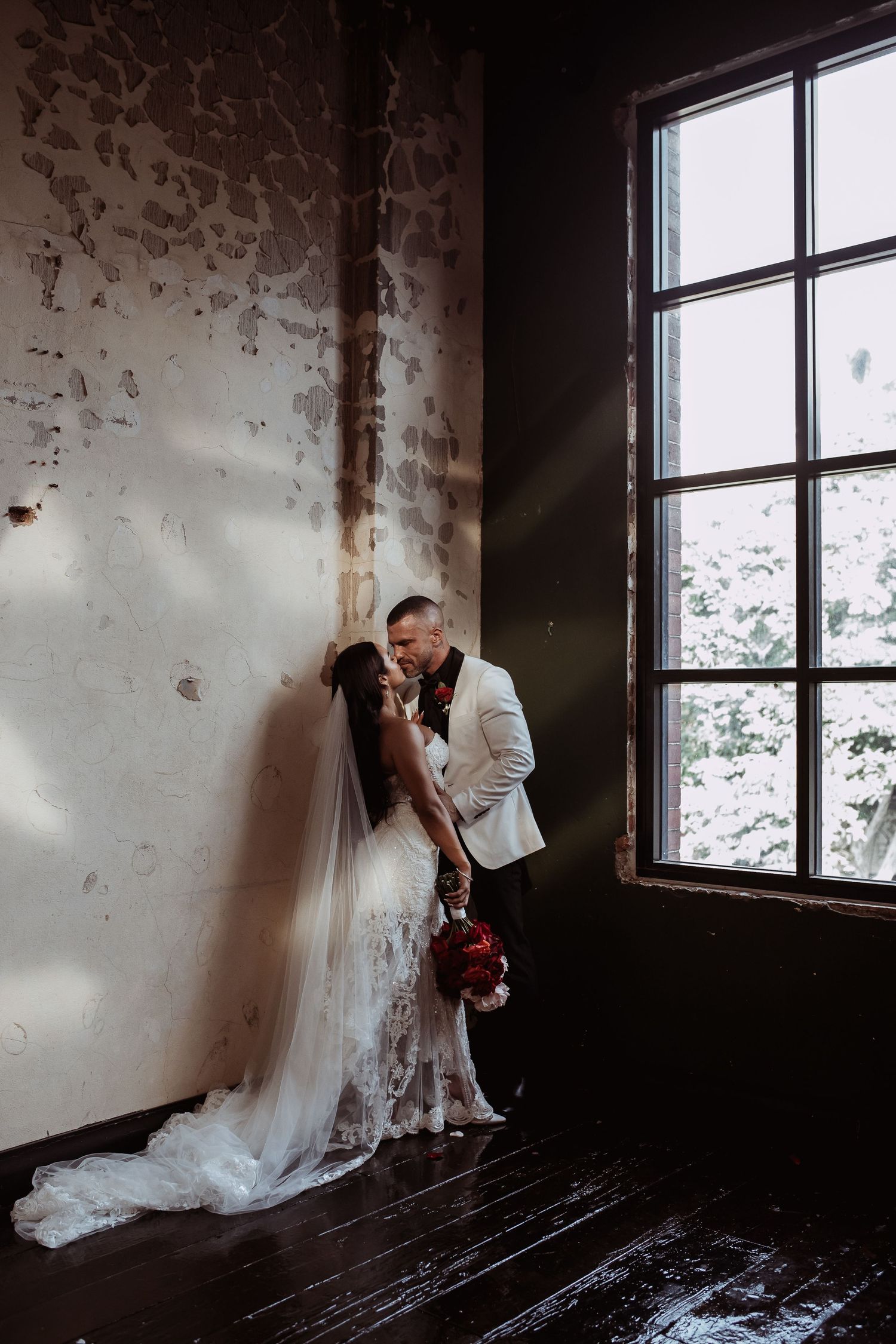 A couple shares an intimate moment near a window in a rustic indoor setting with peeling walls and natural light.