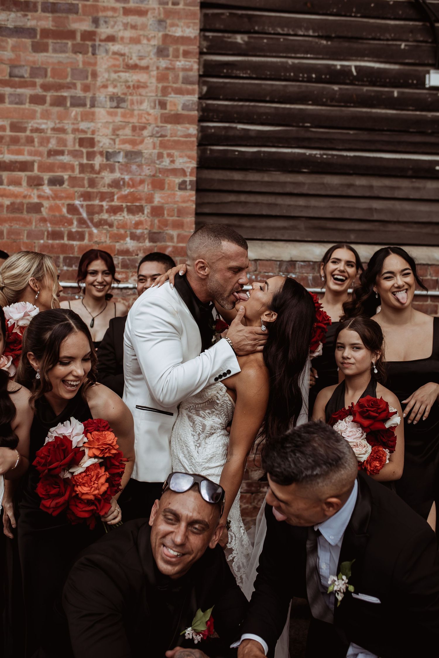 A joyful wedding party laughs together against a rustic brick wall backdrop with red floral bouquets.
