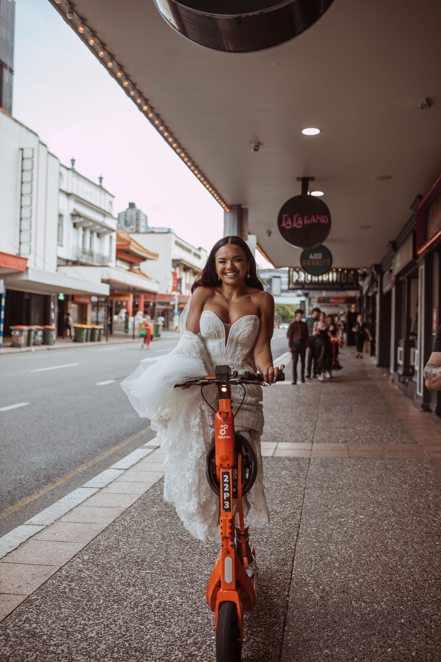 A person in a white wedding dress rides an orange rental scooter down a city sidewalk under covered walkway.