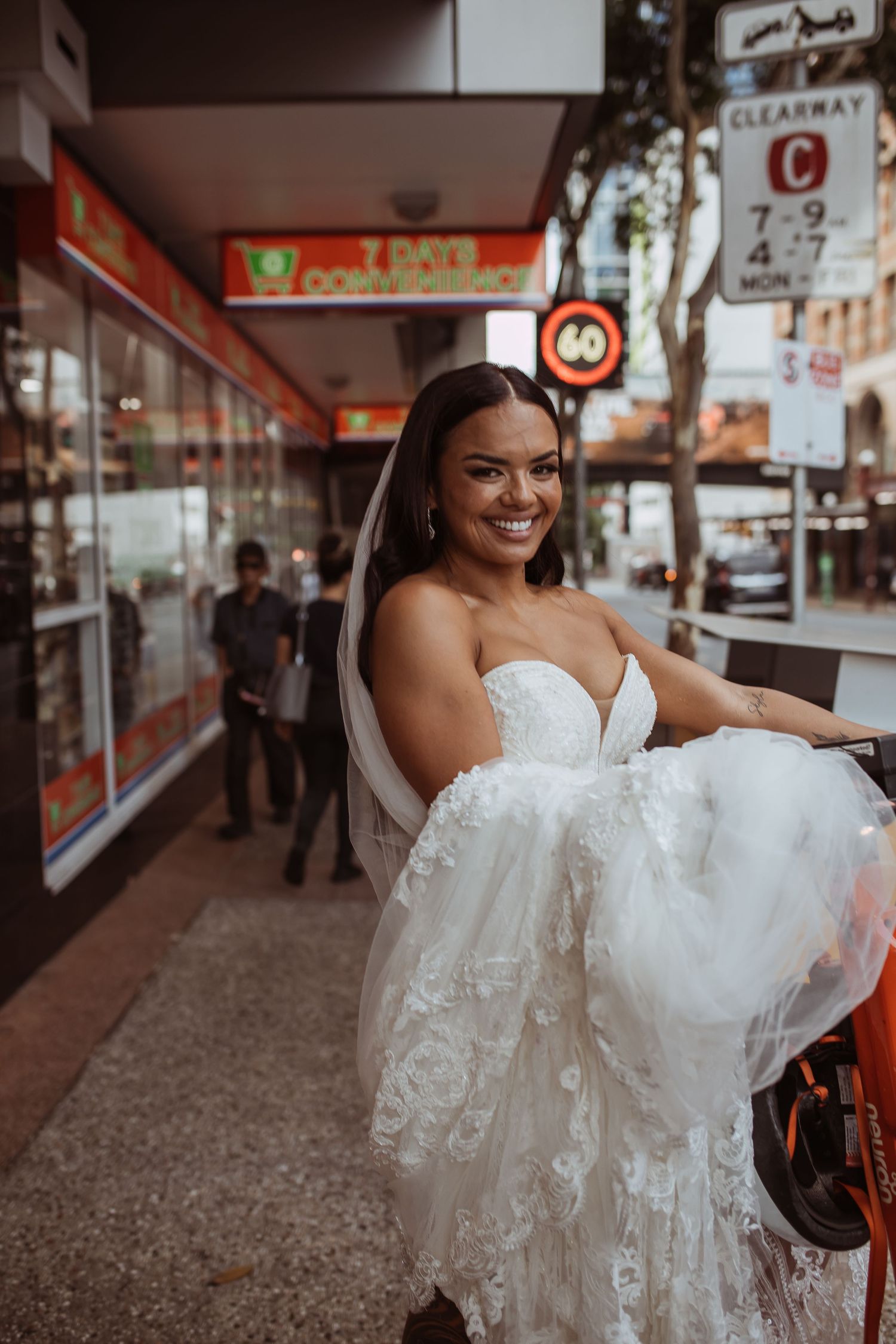A bride wearing a white strapless lace wedding dress smiles while standing on a city street.